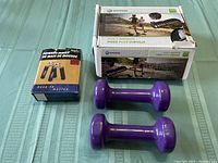 Photo of two purple hand weights, box of Horizon Fitness ankle weights, and box of black foam hand strengtheners on green surface.