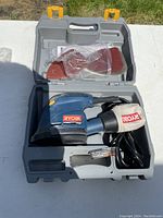 Open gray molded plastic carrying case holding the Ryobi corner cat sander with attached dust collection bag, a few sanding pads in sealed plastic bags, and instruction manual.