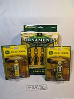 Front view of all three packages: two individual 1950s style John Deere gas pump ornaments and one box of 1920s style ornaments in sealed packaging.