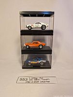 Three stacked display cases each containing one Hot Wheels Camaro car (white, orange, blue) shown from front and side, with handwritten label below.