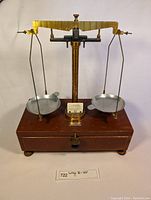 Front view showing brass beam, two metal pans suspended by wires, wooden base with drawer, brass knob, and small label in front.