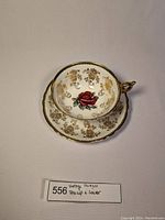 Teacup and saucer set shown from angled side view, displaying red rose and gold floral designs with gold trim