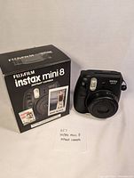 Black Fujifilm Instax Mini 8 instant camera with original box, front angled view showing flash and lens.