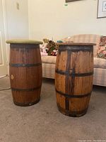Two wooden barrel end tables standing upright on the floor beside a couch. One has a green cushioned top, the other has a wooden top. Shows wood grain and metal bands.
