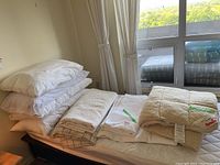 View of stacked white pillows, folded bedspreads, and a quilted mattress pad with intricate stitching on a bed.