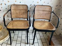 Photo of two wood chairs with black-painted frames and cane seats and backs, showing cane damage needing repair, on a tiled floor with patterned wallpaper.