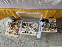 Full wide-angle view of the lot showing three containers of various clip-on earrings and brooches including some vintage pieces on a carpeted floor.