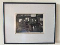 Framed portrait print in simple black frame and white mat showing exterior of brick house with figure on stairs.