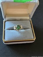 Ring resting in gray jewelry box, front view showing green center and clear accents