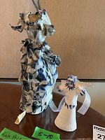 Two pottery figures, a larger 10-inch cream and blue floral design figure and a smaller 5-inch figure with ribbons, showing the broken wing on the smaller figure.