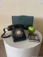 Photo showing the rotary phone, ceramic apple, glass tray, and the mah jong case placed together on a white round pedestal.