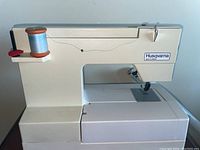 Husqvarna sewing machine with blue thread on spool pin, viewed from front and side showing machine body and needle area.