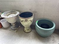 Three garden containers: large metal urn on left with scalloped edge and weathered paint, a smaller metal urn with ornamental decorations in middle, and a green glazed ceramic rounded pot on right.