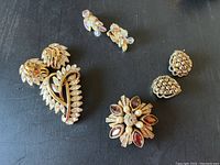Group photo of all vintage costume jewelry including Trifari brooch and earrings, DeNicola brooch, Weiss and Kramer clip-on earrings on black surface.
