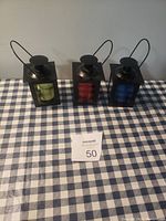 Front view of three black metal lantern candle holders with green, red, and blue glass panels, placed on blue and white checkered cloth.