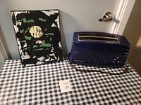 Image shows a blue KitchenAid toaster and a small framed abstract art picture on a blue and white checkered tablecloth in front of a gray door.