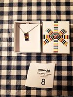 Photo of the brand-new Hudson necklace in its gift box featuring a colorful striped pendant on a silver-tone chain alongside the box lid with a matching striped ribbon design and a MaxSold tag.