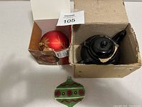 Box containing black ceramic tea pot, red Christmas ball ornament in original box, and green Christmas ornament placed next to boxes.