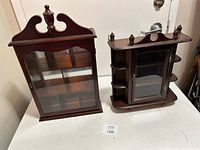 Photo showing two wooden wall hanging cabinets on a white table, one with a glass front and simple design, the other with mirrored back, side shelves, and finials on top.