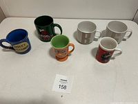 Front view of all six mugs lined in two rows on a table showing colors and inscriptions clearly