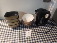 Photo showing one black electric kettle, three ceramic bowls stacked inside each other, and four rectangular beige dishes stacked.