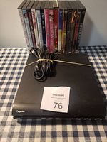 Photo showing black RCA DVD player with power and AV cables wrapped on top, along with 12 assorted DVDs standing upright behind it on a checkered cloth.