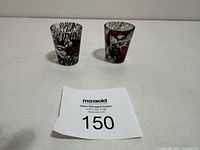 Two Harley Quinn shot glasses, clear with colorful character designs, displayed on a white surface.