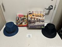 Two fedora-style hats, two board games (The Godfather and Clue), and a puzzle with horse carriage scene lined up on a white surface.