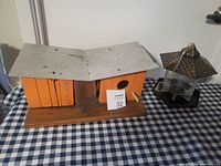 Photo displaying a homemade wooden bird house next to a bird feeder, both resting on a checkered cloth surface.