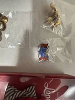 Three pairs of earrings in clear plastic bags on a white surface with a partial view of a leopard print jewelry box lid with red interior.