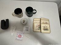 Photo showing two ceramic mugs, six floral square coasters, two white heart-shaped decorative dishes, a black U-shaped item, a white heart-shaped ornament, a pink magnet, and a clear plastic clip on a white surface.