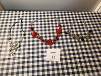 Three necklaces on blue and white checkered tablecloth: one sterling silver with coin charms, one red beaded, one silver with dark stone charms
