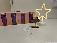 Photo showing boxed set of four purple glasses with gold rims, star-shaped neon light, and small metallic Vegas sign spaced in front.