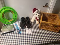 Photo showing wicker basket, stuffed dog toy, two green toy frisbees, black sneakers, and dog collars on a blue and white checkered tablecloth.