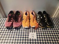 Three pairs of women's athletic sneakers lined up on a table with a lot number card in front: pink New Balance, orange Nike, and black adidas sneakers.