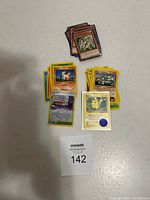 Top-down photo showing two stacks of cards on a gray surface: a small stack of brown-bordered Yu-Gi-Oh cards and a larger stack of yellow-bordered Pokemon cards including a Pikachu card with a blue circular stamp.