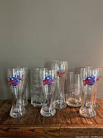 Front view of all 7 beer glasses including Planet Hollywood and beer mugs on wood surface, against gray wall