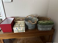 Seven assorted containers including fabric-covered box, wooden-look box with starfish, metal baskets with rope handles, wicker basket, and floral foam container displayed on wooden surface.