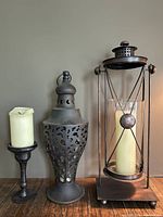 Three decorative candle holders arranged side by side: a heavy metal pedestal holder with candle, a metal urn-style holder with open cut-out design, and a tall metal and glass lantern-style gimble holder with a candle inside.