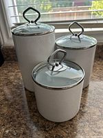 Photo showing three white kitchen cannisters with glass lids and metal ring handles, varying heights, on a countertop near a window.