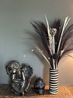 Cherub statue, small urn, and tall wicker vase with dried decorations on a wooden surface against a plain wall.