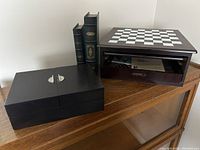 Overall view of wooden multi-game box with chessboard top, two decorative book boxes, and black box with handles on wooden shelf