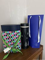 Photo showing all three items: a large patterned cylindrical gift box with green ribbon handles, black faux leather bottle holder, and blue cardboard gift bag