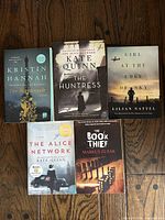 Top view of five WWII-themed fiction paperback books placed on wooden floor, titles visible: 'The Nightingale', 'The Huntress', 'Girl at the Edge of the Sky', 'The Alice Network', and 'The Book Thief'.