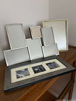 Four picture frames arranged on a wooden surface: a multi-photo nickel frame holding six photos, a brass-colored 8x10 frame, a silver frame holding three 3x3 photos, and a wooden multi-photo frame.