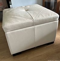 Side view of the square bonded leather ottoman showing beige leather upholstery and black feet.