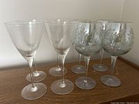 Eight wine glasses arranged in two groups: four clear triangular shaped glasses in front and four etched glasses with floral patterns behind.