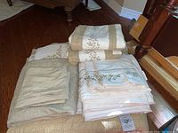 Full view of folded beige and white quilt with floral embroidery and stacked white sheets and pillowcases with similar embroidery on wooden floor