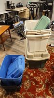 Photo of three large white plastic baskets stacked, green lids visible, surrounded by various plastic bins of multiple colors placed on a carpeted floor.