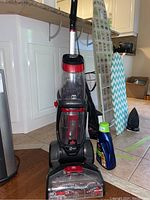 Full view of the Bissell ProHeat Revolution 2X upright carpet cleaner with red and black design, placed on tile floor beside ironing board and cleaning solution bottle.